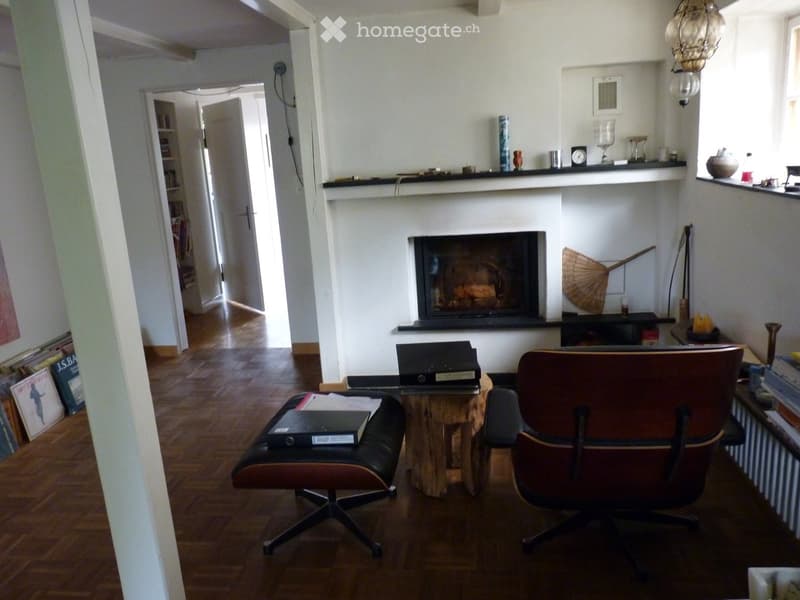 Living room with wooden flooring, fireplace, two chairs, small table, and shelves with decorative items.