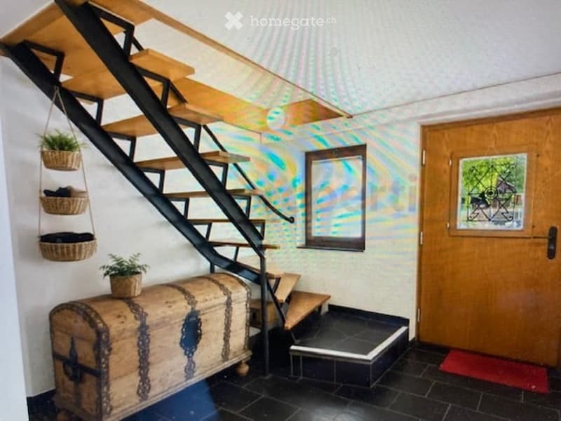 Staircase with wooden steps and black railing, colorful wall art, wicker baskets with plants, wooden chest at the bottom, and a wooden door with glass panel.