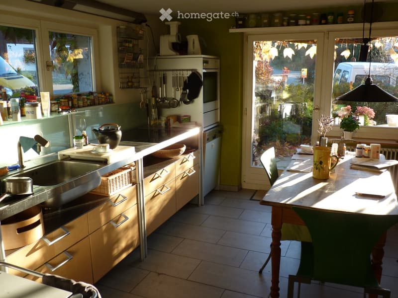 Bright kitchen with wooden cabinets, stainless steel sink, and a dining area with a table and chairs. The kitchen features various utensils, a teapot, and a refrigerator.