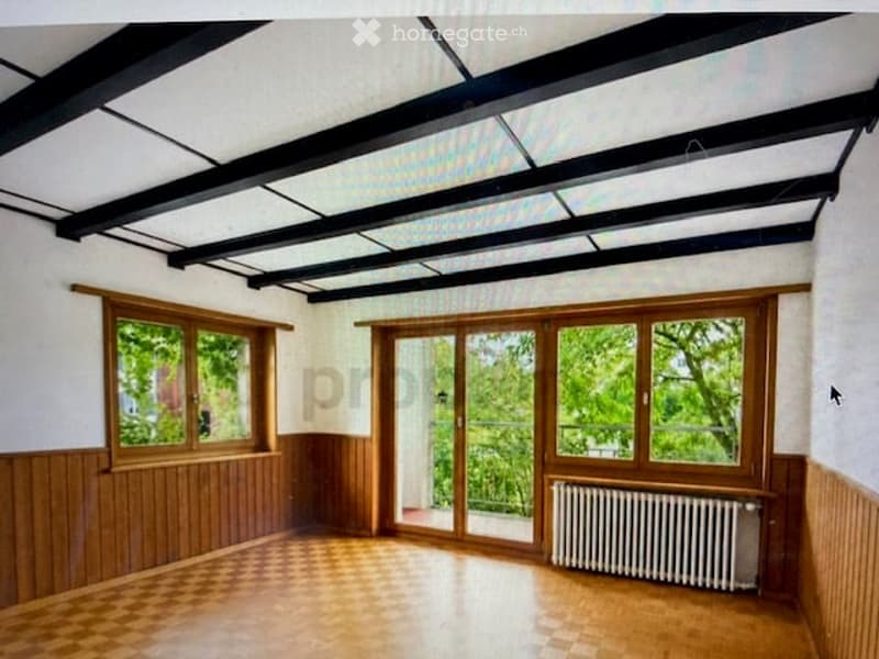 Empty room with wooden flooring, white walls, large windows, and a radiator. The room features exposed wooden beams on the ceiling and a sliding glass door leading to a balcony.