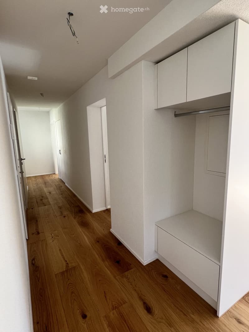 Empty hallway with light wooden flooring, white walls, built-in white closet with shelves and drawers, and several doors leading to other rooms.