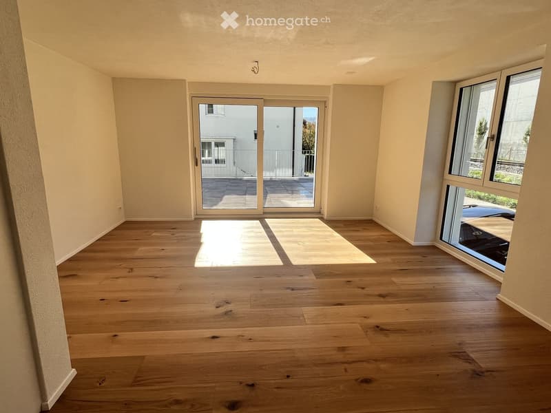 Empty room with light wooden flooring, large windows, and sliding glass doors leading to a balcony.