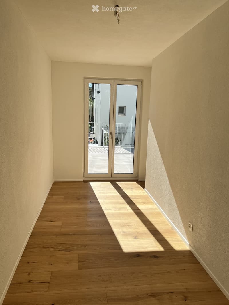 Empty hallway with light wooden flooring and beige walls, featuring a large glass sliding door leading to a balcony area.