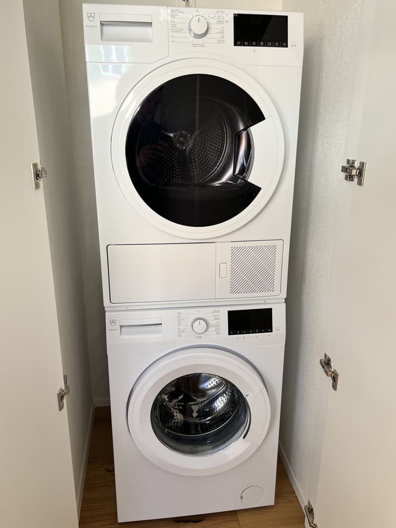 White washing machine and dryer unit installed in a small space with wooden flooring and white walls.