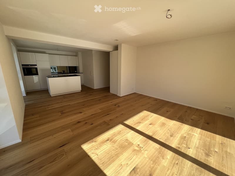 Empty room with wooden flooring and white walls, featuring an open kitchen area with cabinets and appliances visible in the background.