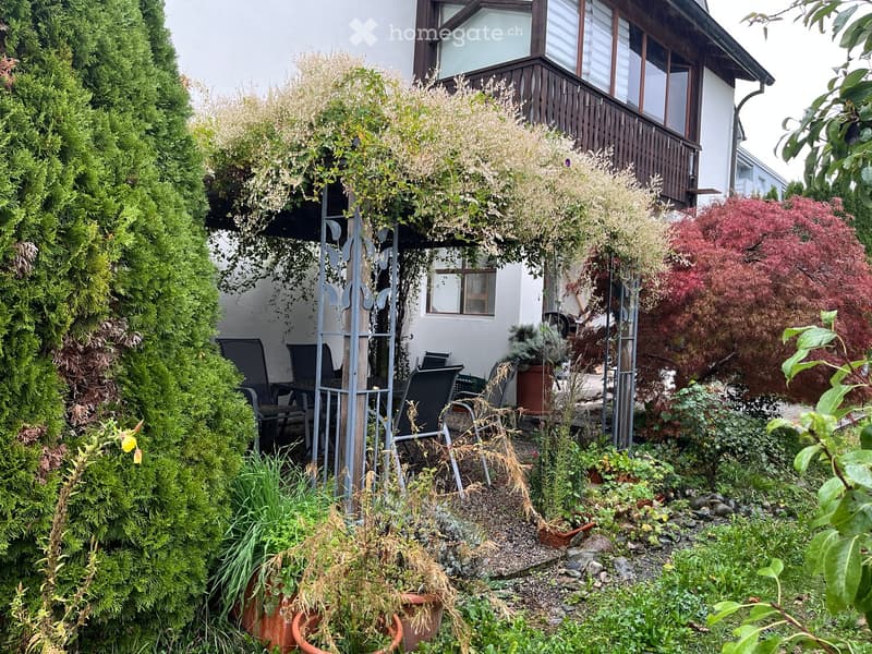 Garden area with a covered seating area, surrounded by lush greenery, including shrubs, potted plants, and a small table with chairs.