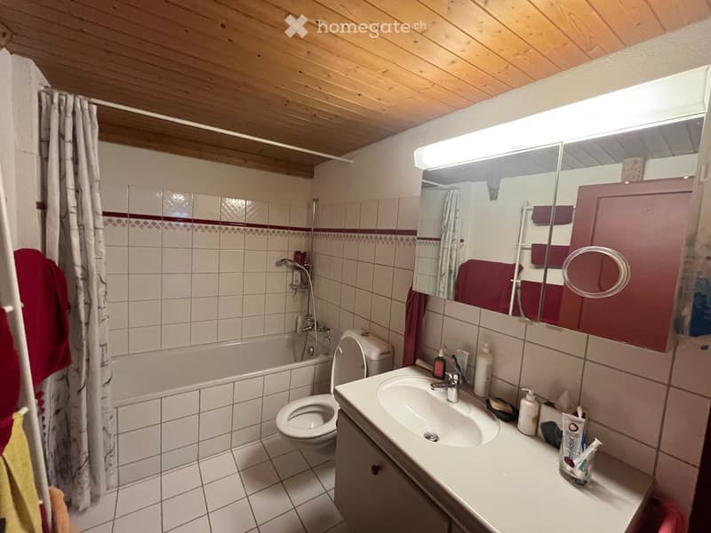 Bathroom with white tiled walls and floor, a sink with a mirror above it, a toilet, a bathtub with a shower curtain, and various toiletries on the sink counter.