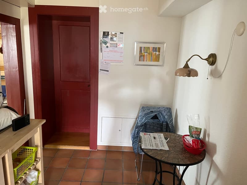 A small, furnished room with a round table, a chair draped with a blue cloth, a wall-mounted lamp, a mirror, a framed picture, and a newspaper on the table. A green basket and a wooden shelf are visible on the left side.
