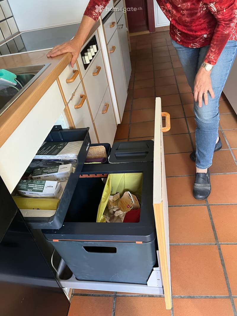 A kitchen with white cabinets, wooden handles, and a trash bin with multiple compartments. A person is seen interacting with the cabinets.