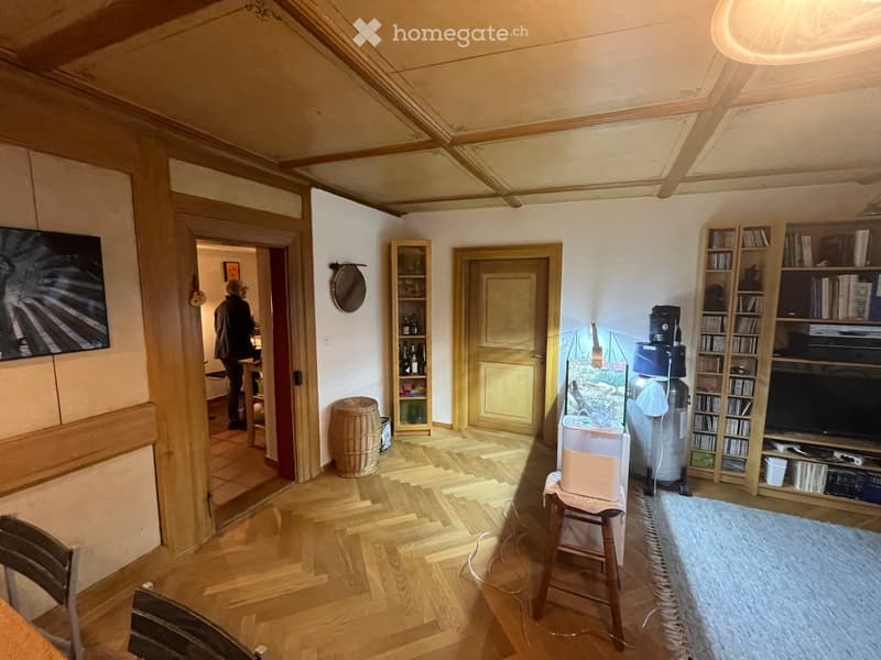 A furnished room with wooden flooring, a large shelf unit, a small table, a mirror on the wall, and a screen. There is a doorway leading to another room.