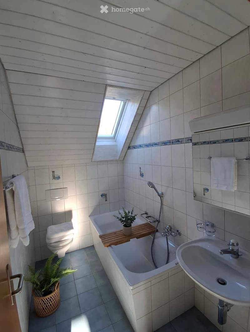Bright, modern bathroom with white tiled walls and wooden slats on the ceiling. Features a bathtub with a wooden tray, a toilet, a sink with a mirror, and a small potted plant near the door.