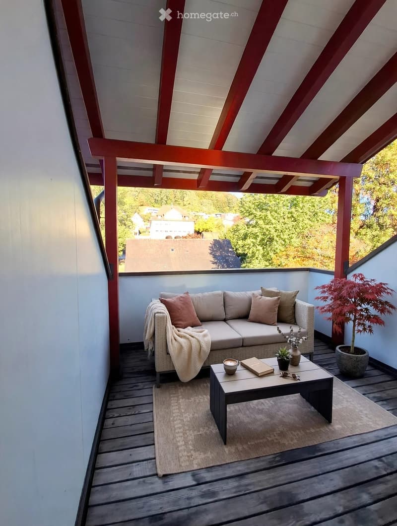A cozy balcony with a wicker sofa, coffee table, small plant, and decorative items, featuring wooden decking and a slatted roof.