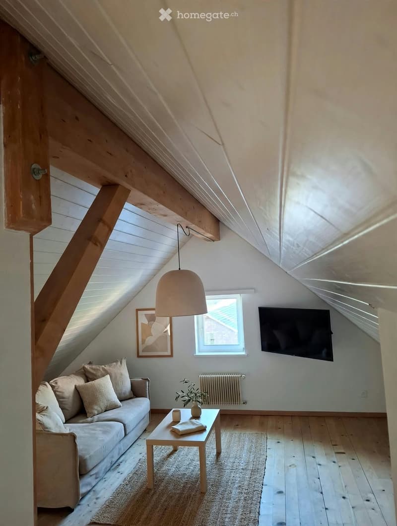 Cozy attic living room with slanted wooden ceilings, large window, sofa, coffee table, table lamp, television, and decorative items.