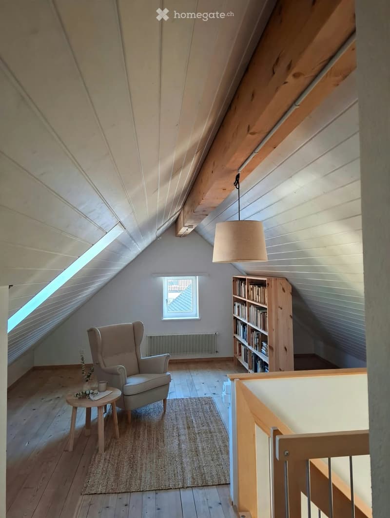 Cozy attic room with wooden floors, slanted ceilings, a large window, a comfortable armchair, a small side table with a cup, a bookshelf filled with books, and a hanging lamp.