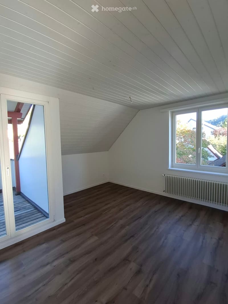 Empty attic room with wooden flooring, white walls, and a slanted ceiling. Features a large window and a radiator on one wall.