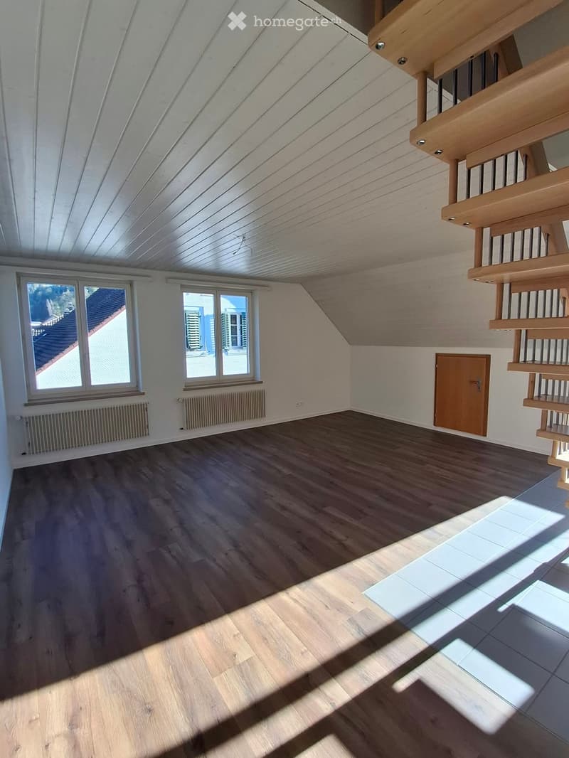 Empty attic space with wooden flooring, white walls, exposed wooden beams, and large windows offering a view of the roof and surrounding area.
