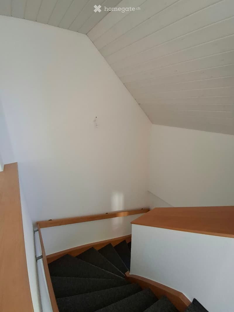 A narrow staircase with wooden handrails and dark carpeted steps, leading upwards with white walls and a slanted ceiling.
