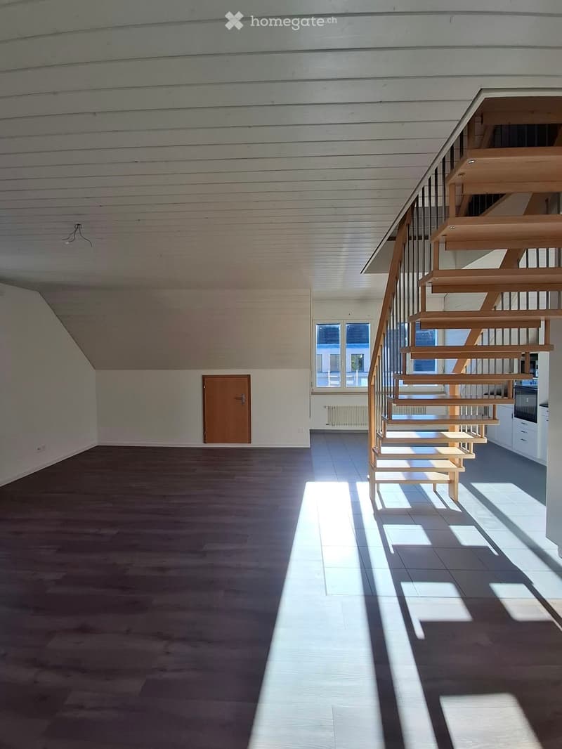 Empty room with wooden floor, wooden staircase, and large windows letting in natural light.