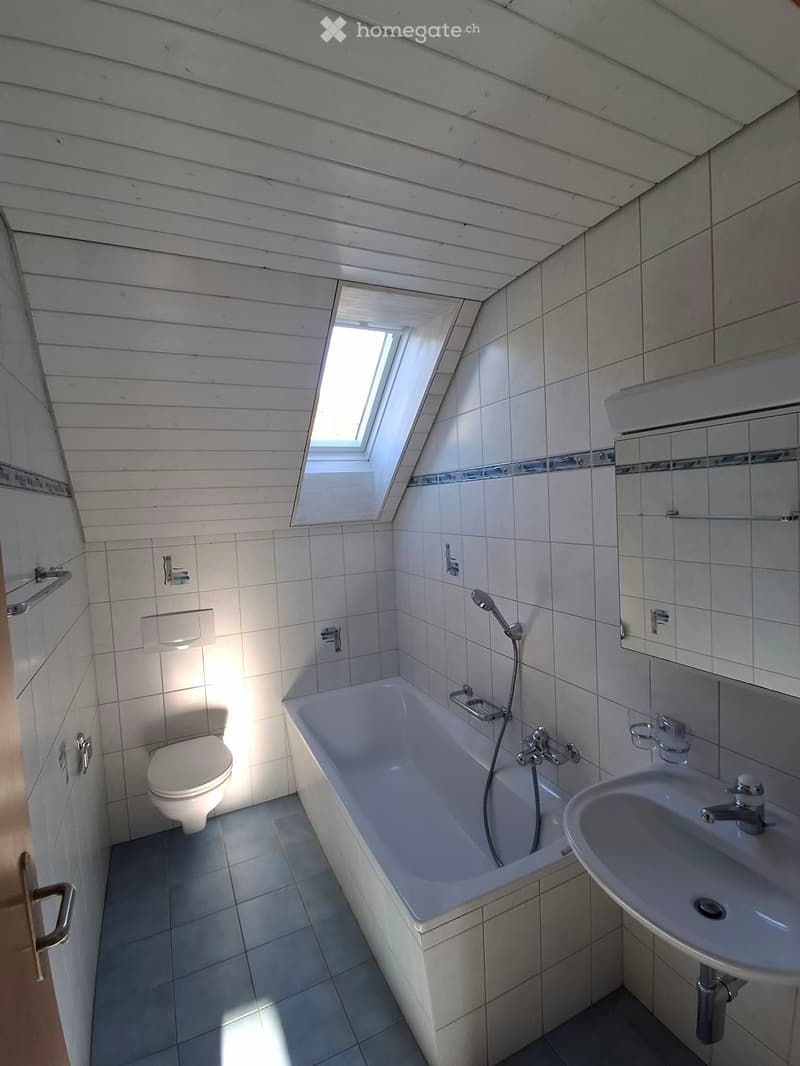 Modern bathroom with a bathtub, toilet, sink, and showerhead. Features white tiled walls, a skylight window, and a wooden slanted ceiling.