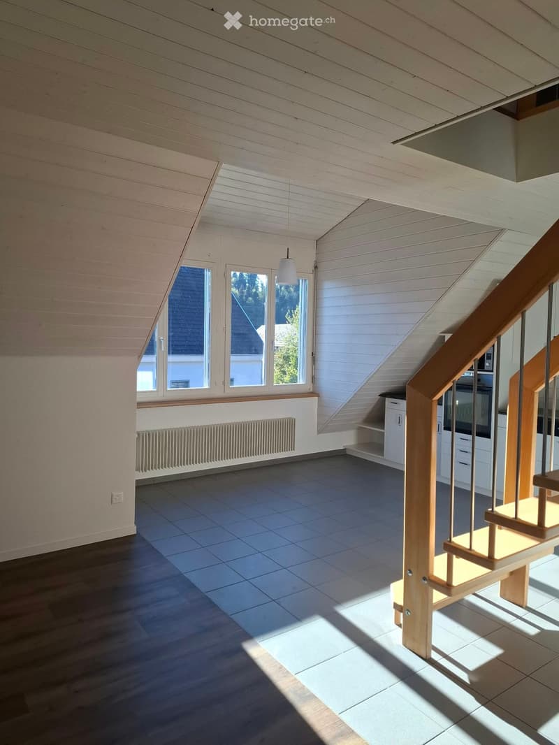 Bright, empty attic space with wooden beams, large windows, and a staircase leading upwards. The floor is tiled, and the walls are painted white.