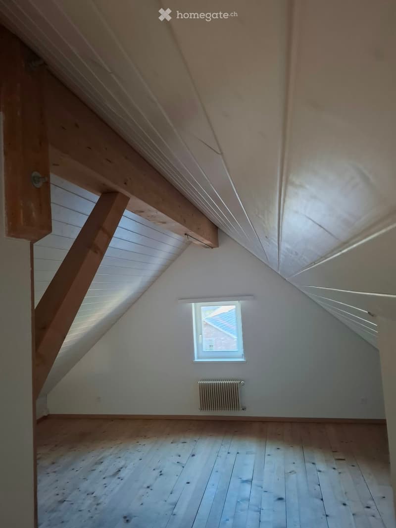 Empty attic room with wooden floor, slanted wooden ceiling, single window, and radiator.