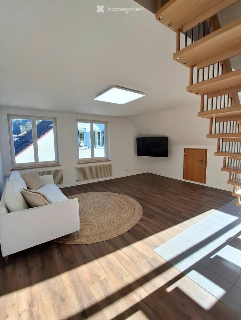 Modern living room with white walls, wooden flooring, a white sofa, a round jute rug, a flat-screen TV mounted on the wall, and large windows offering a view of the roof.