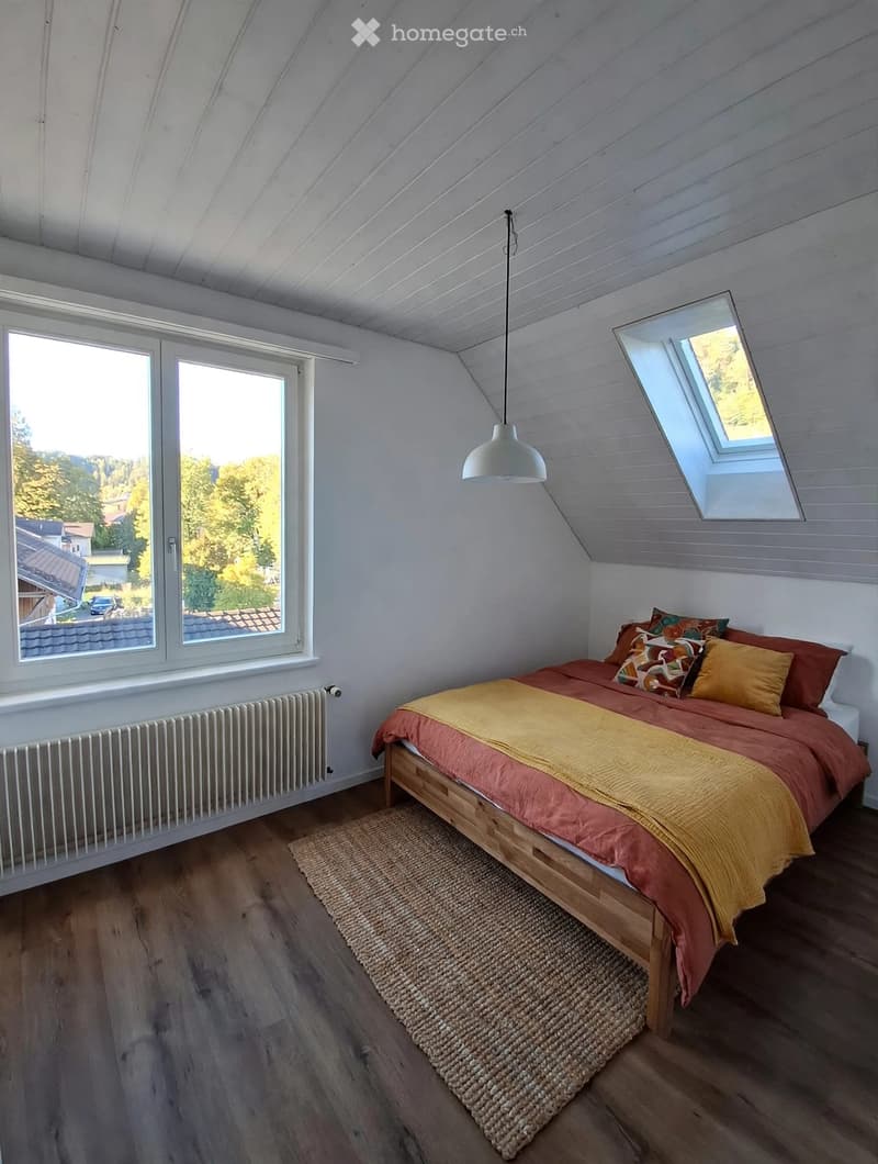 Bright bedroom with wooden floors, large windows, skylight, bed with colorful bedding, and a woven rug.