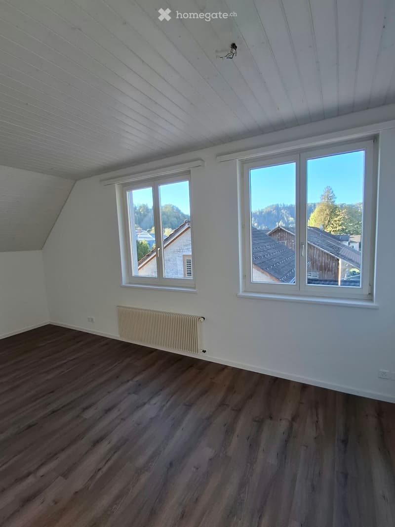 Empty attic room with wooden flooring, white walls, large windows, and a radiator. No furniture or equipment present.