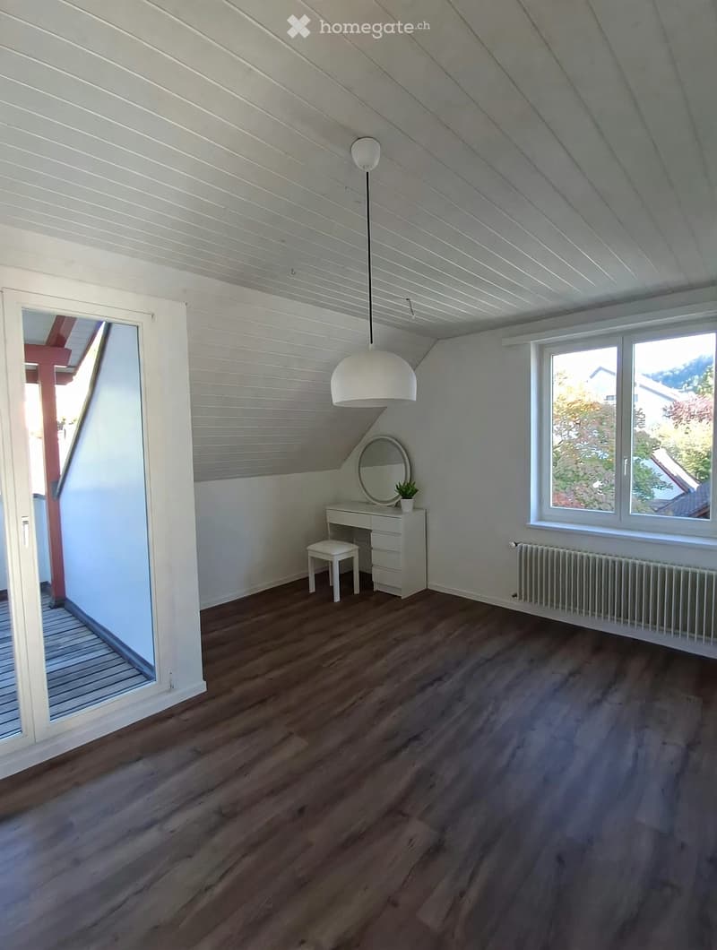 Bright attic room with wooden flooring, white walls, and a slanted ceiling. Features a small desk with a mirror, a stool, a pendant light, and a radiator.