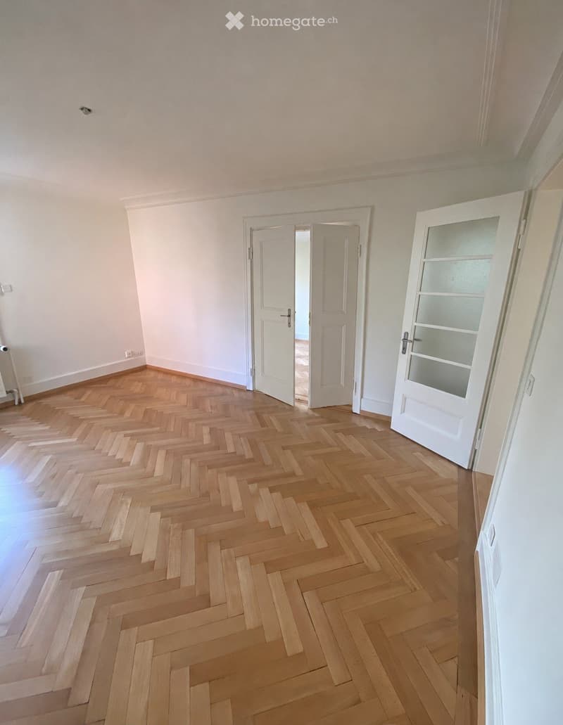 Empty room with herringbone-patterned wooden flooring, white walls, and two open doors leading to other spaces.