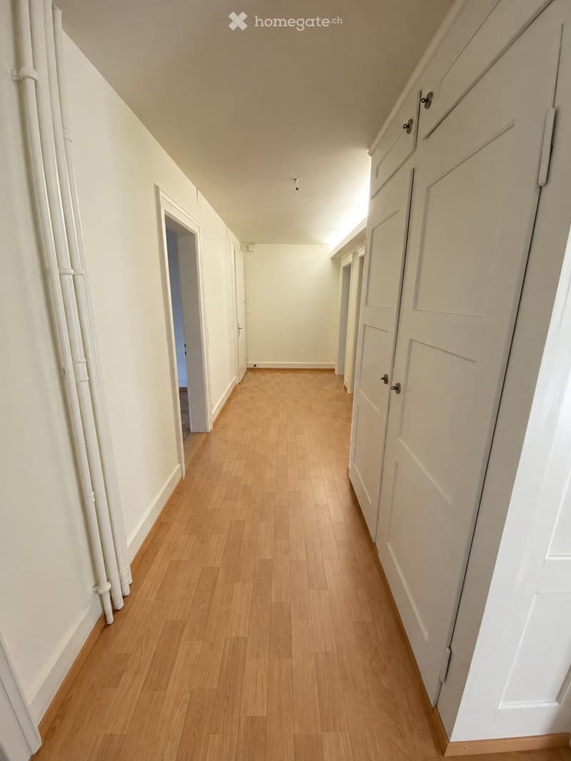A narrow hallway with light wooden flooring, white walls, and several closed doors on both sides. The ceiling is sloped, and the lighting is bright, creating a clean and minimalist look.