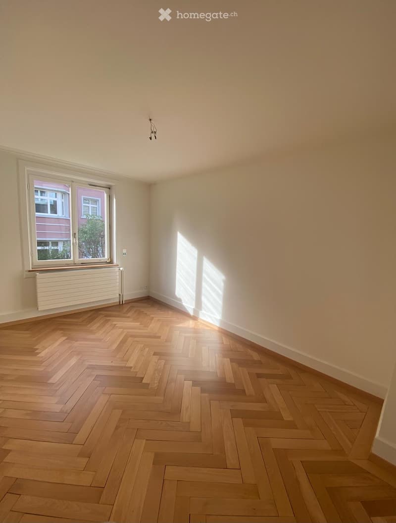Empty room with wooden herringbone flooring, white walls, and white radiator. Sunlight streaming in from the window.