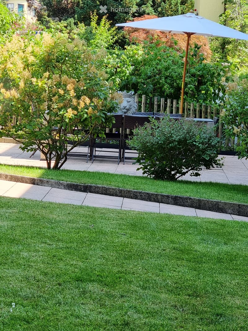 Garden area with grassy lawn, shrubs, outdoor table, chairs, and umbrella, decorative lion statue, white fences, trees, bushes, and plants
