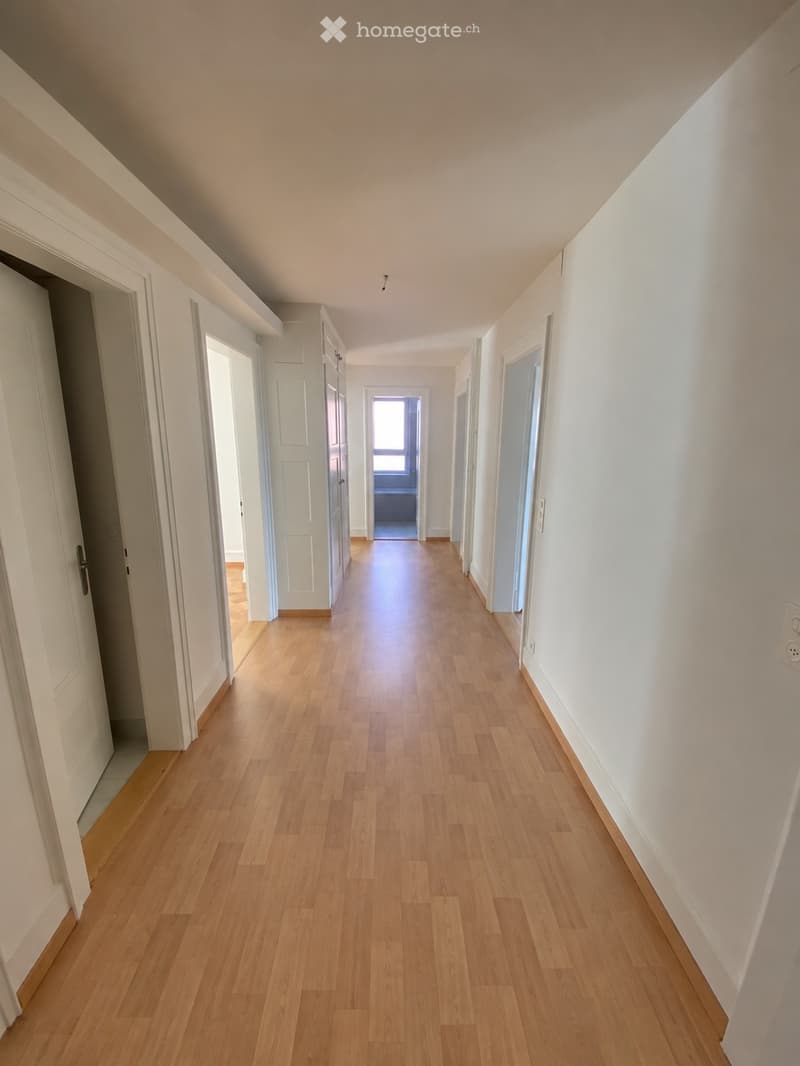 A long, well-lit hallway with wooden flooring and white walls, featuring multiple doors on both sides and a window at the far end.