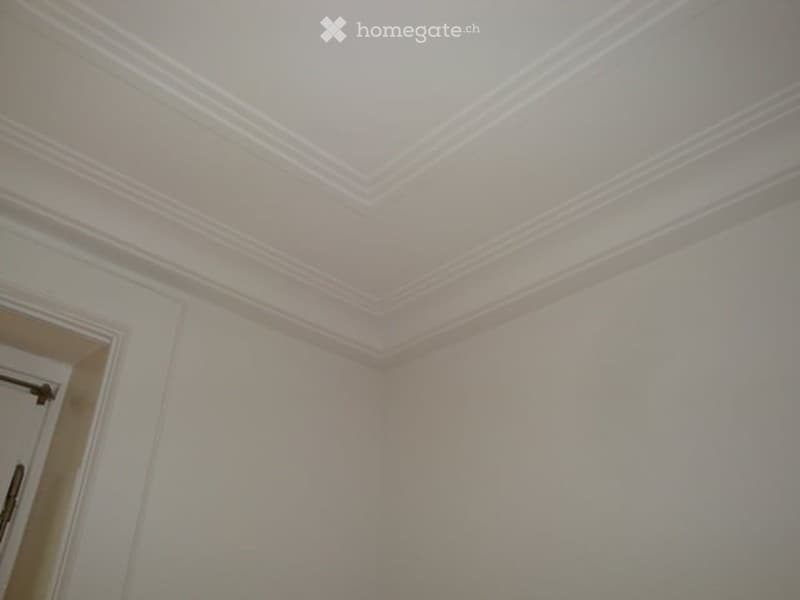 The image shows the interior of a room with white walls and a decorative crown molding on the ceiling. The room appears to be a living room or bedroom with a simple, minimalist design.