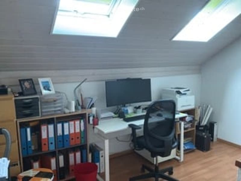 A modern, well-lit home office with wooden flooring, white walls, and skylights. The room features a white desk with a computer monitor, a black office chair, a printer, a bookshelf filled with binders, and a small storage cabinet.