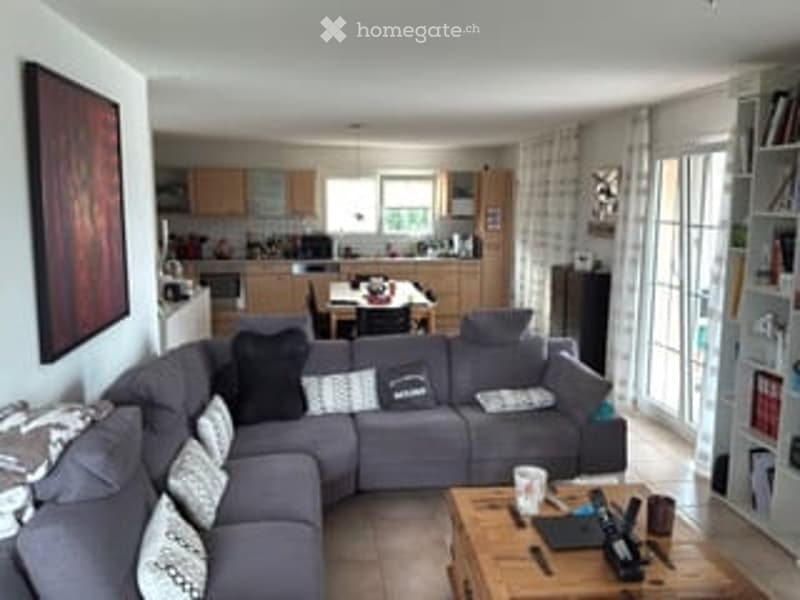 Open-plan living and dining area with a grey sectional sofa, coffee table, dining table with chairs, and a modern kitchen in the background.