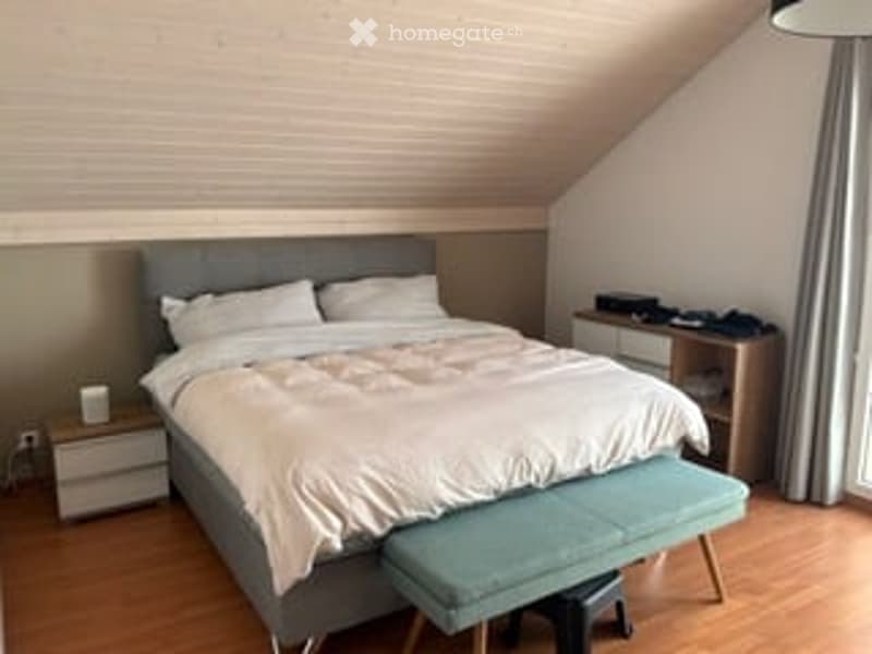 Cozy bedroom with a large bed, nightstand, bench, desk, and chair. Light wood flooring and soft lighting.