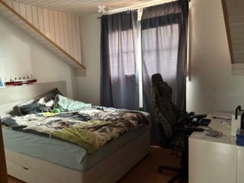 Bedroom with a bed, nightstand, chair, and curtains. The bed is unmade, and there is a small table with items on it.