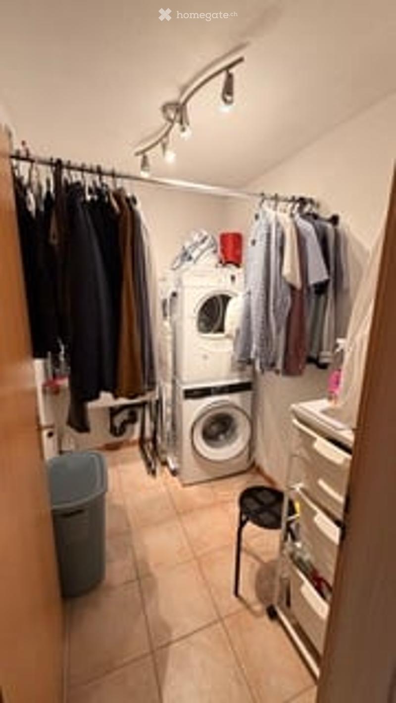 Laundry room with a washing machine, a small stool, a wardrobe with hanging clothes, and a light fixture on the ceiling.