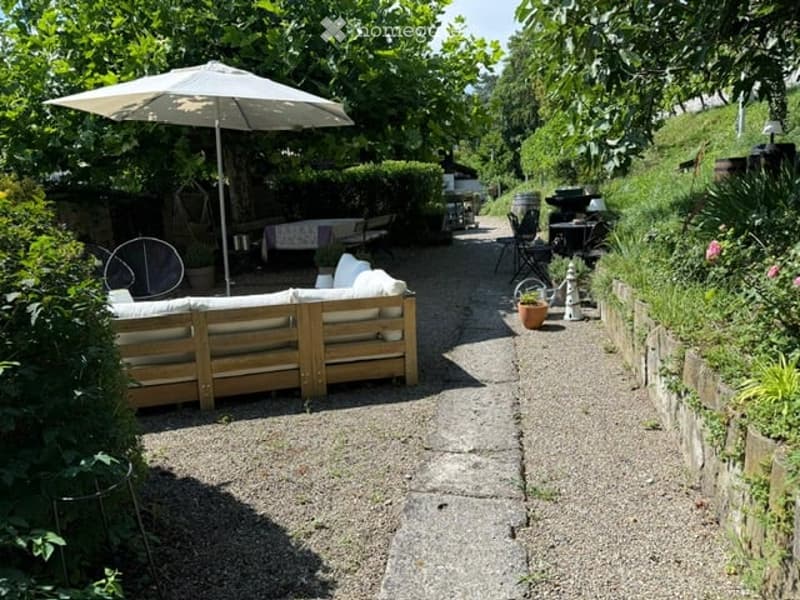 A serene garden patio with a wooden sofa, umbrella, and small table surrounded by lush greenery and a stone pathway.
