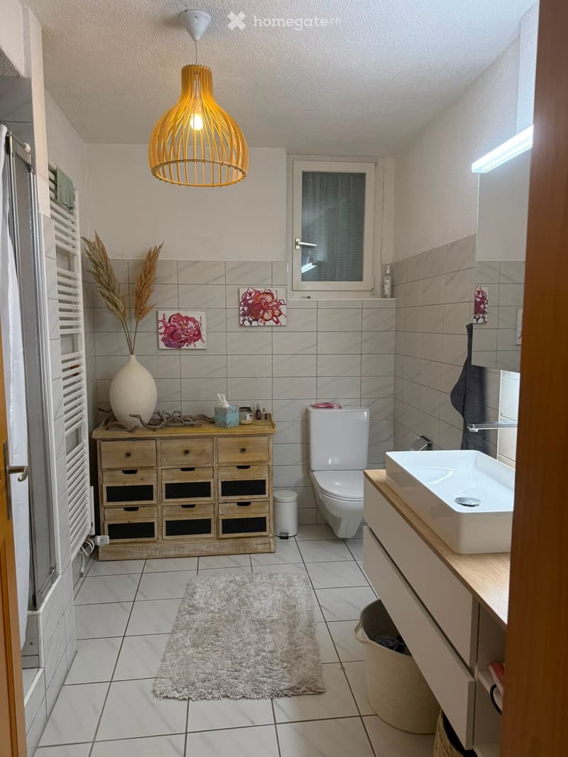 Modern bathroom with white tiles, a toilet, a sink, a wooden cabinet, a white vase with dried plants, a fluffy rug, and a hanging light fixture.