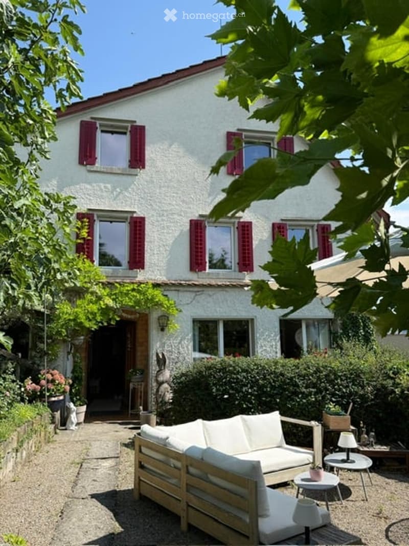 A two-story white house with red shutters and a well-maintained garden. The front features a small seating area with a white sofa and wooden benches.