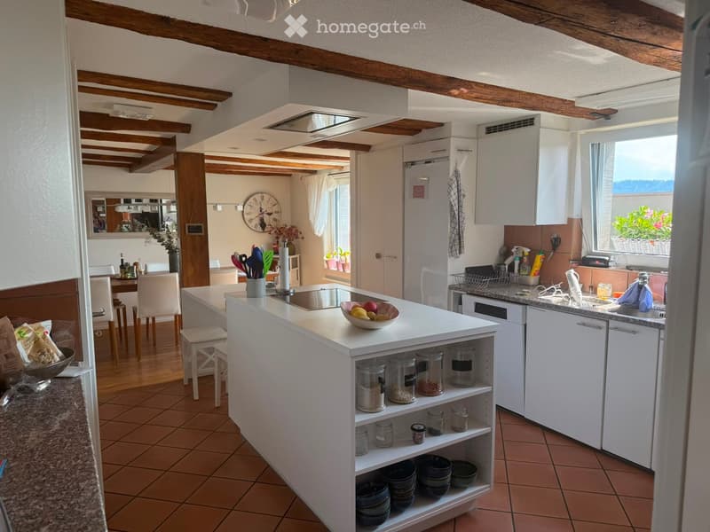 Bright kitchen with white cabinets, a central island, and a dining area. Features include a sink, refrigerator, and various kitchen utensils and items on the counter and shelves.