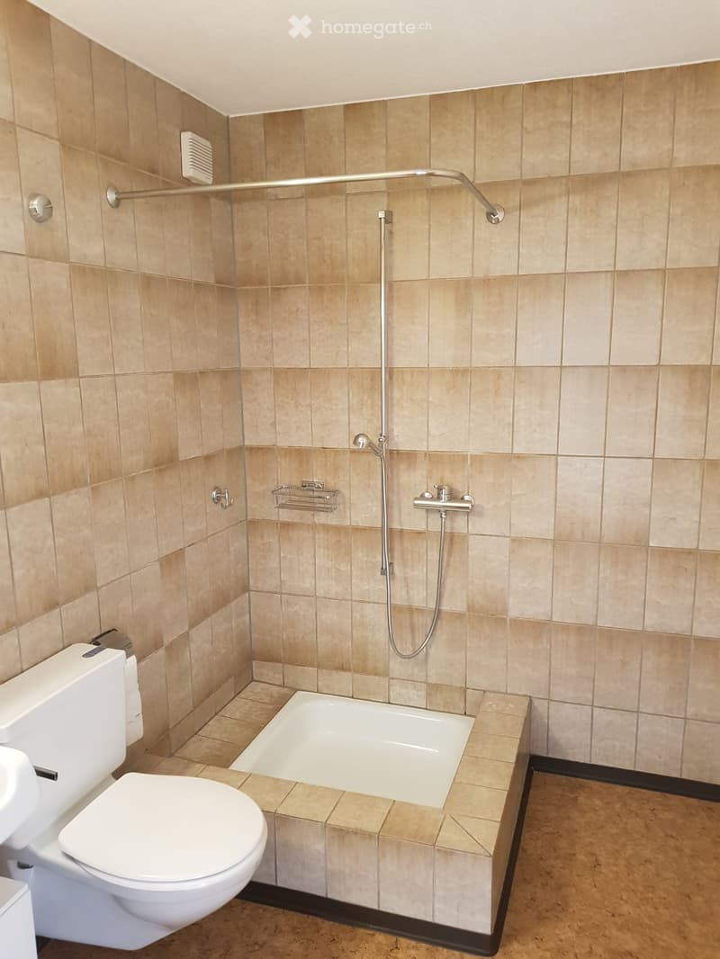 Modern bathroom with beige tiled walls and floor, featuring a bathtub, toilet, showerhead, and a handheld shower rail.