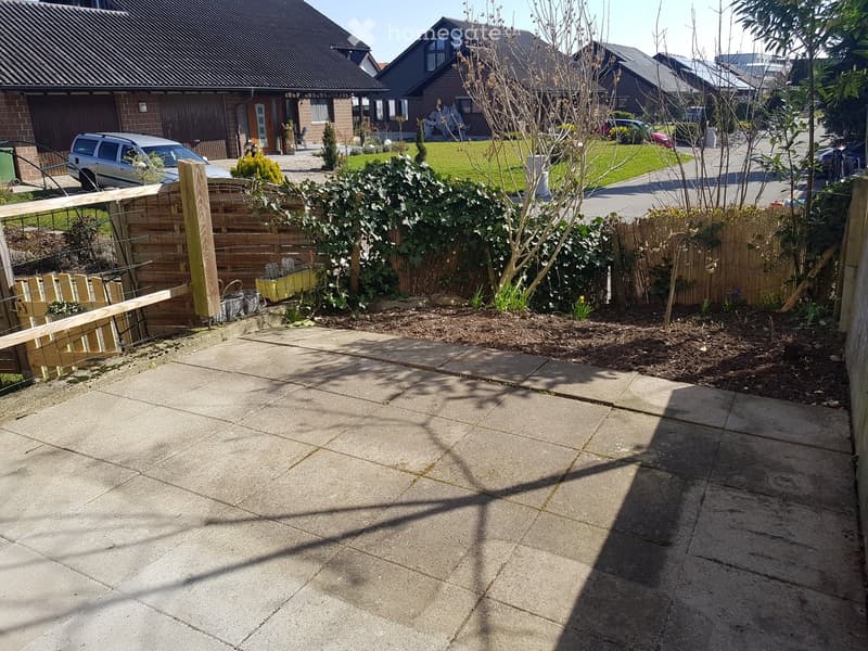 Garden area with a paved concrete patio, wooden fence, and small plants and shrubs. The background shows a residential neighborhood with houses and parked cars.
