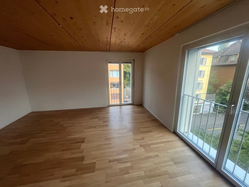 Empty room with wooden flooring, white walls, and large windows offering a view of the street and neighboring buildings.