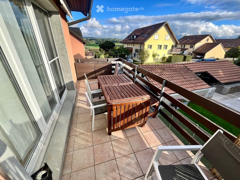 Balcony with tiled floor, wooden table, and chairs, overlooking a residential area with houses and greenery.