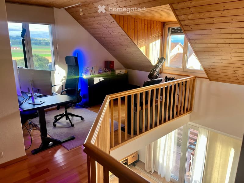 Attic room with wooden slanted ceilings, large windows offering a scenic view, a desk with a chair, a telescope on a stand, and a cabinet.