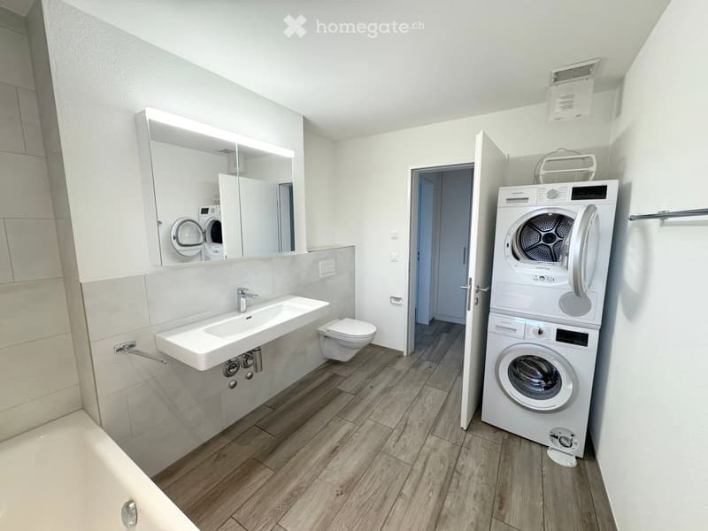 Modern bathroom with a sink, toilet, mirror, and a stacked washer and dryer unit. The space features light-colored tiles and wooden flooring.